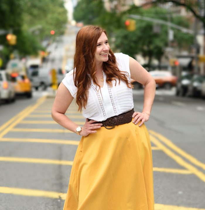 Woman posing in street with hands on hips looking away from camera and smiling while wearing yellow skirt and white shirt